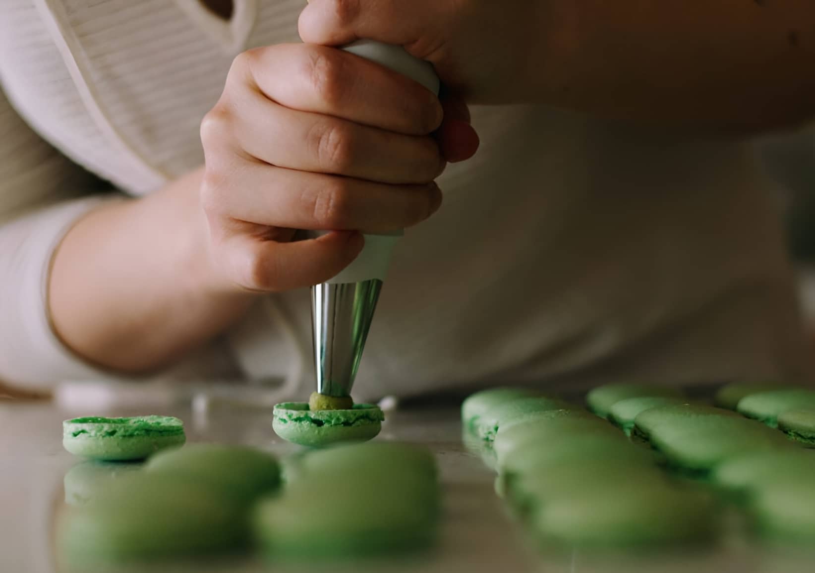 A close up shot of a woman's hands, using a piping bag to squeeze delicious green filling into rows of green macarons